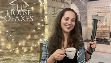 Kate wearing a plaid shirt and holding a cup of tea in one hand and an axe in another in front of a sign reading "the house of axes"